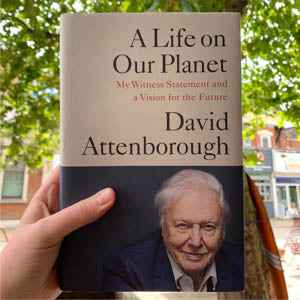 Book cover of "A Life on Our Planet" by David Attenborough held outdoors against a leafy background.