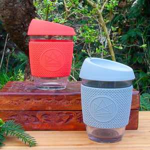 Glass coffee cup by Neon Kactus with red and grey silicone sleeves on a wooden surface.