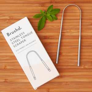 Stainless steel tongue scraper by Brushd on wooden surface with packaging.