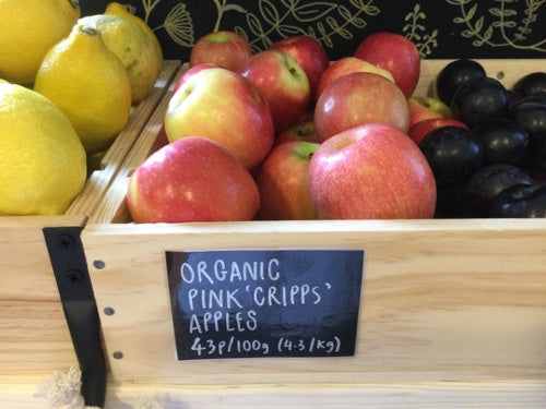 Organic apples on display in a wooden crate, labeled "Organic Pink Cripps Apples" with price and weight details.